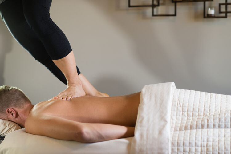 A massage therapist massaging a male client's back with her feet.